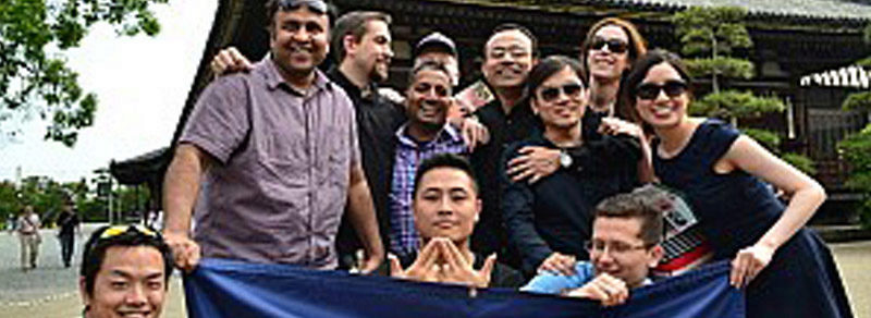 A group of people posing together outdoors, smiling and holding a blue banner, in front of a traditional structure with greenery surrounding it.