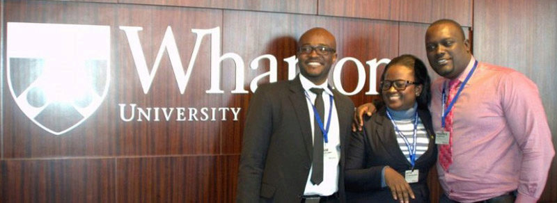 Three people standing and smiling in front of a wall with the Wharton University logo.